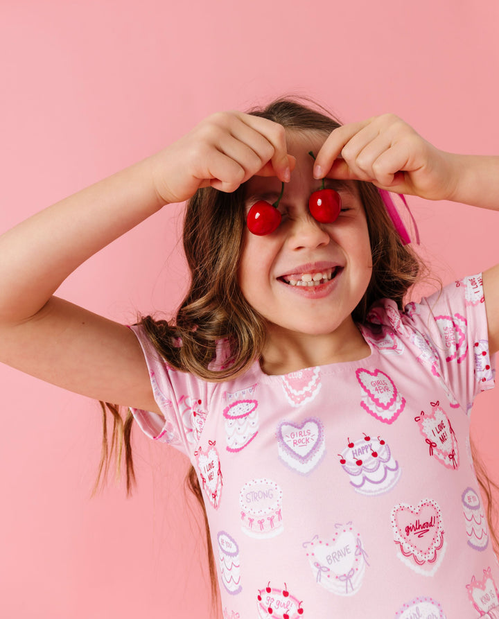 young girl holding cherries in light pink square neck twirl dress with heart shaped cake print featuring words of affirmation for Valentine's Day