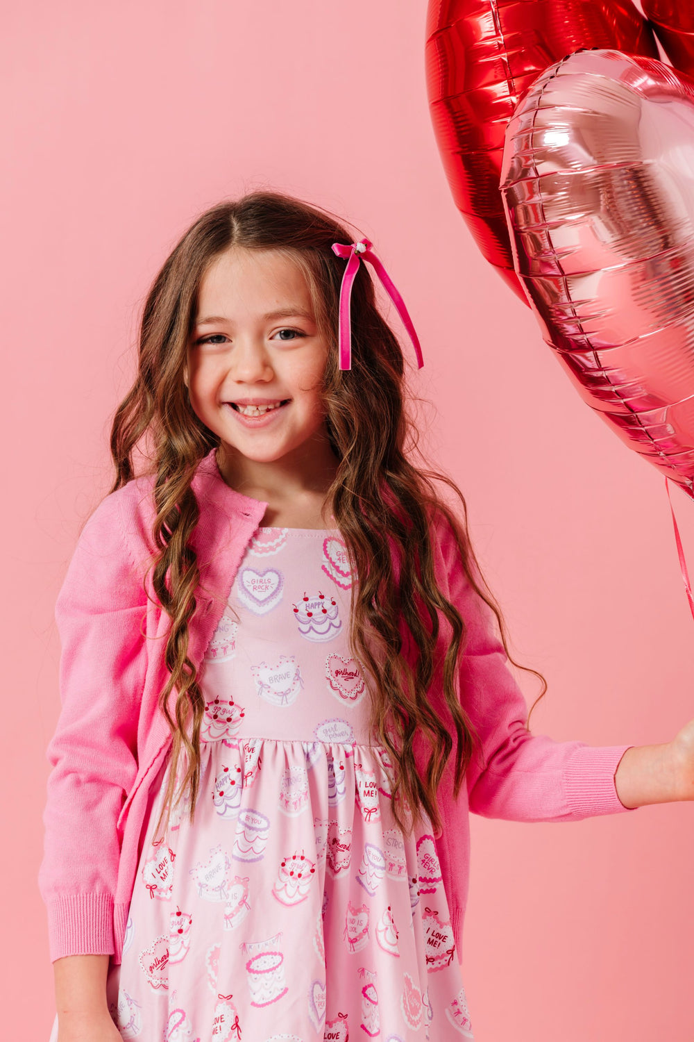 girl holding heart balloons in pink valentines themed dress with cakes and words of affirmation paired with pink cardigan and velvet ribbon rosette bow