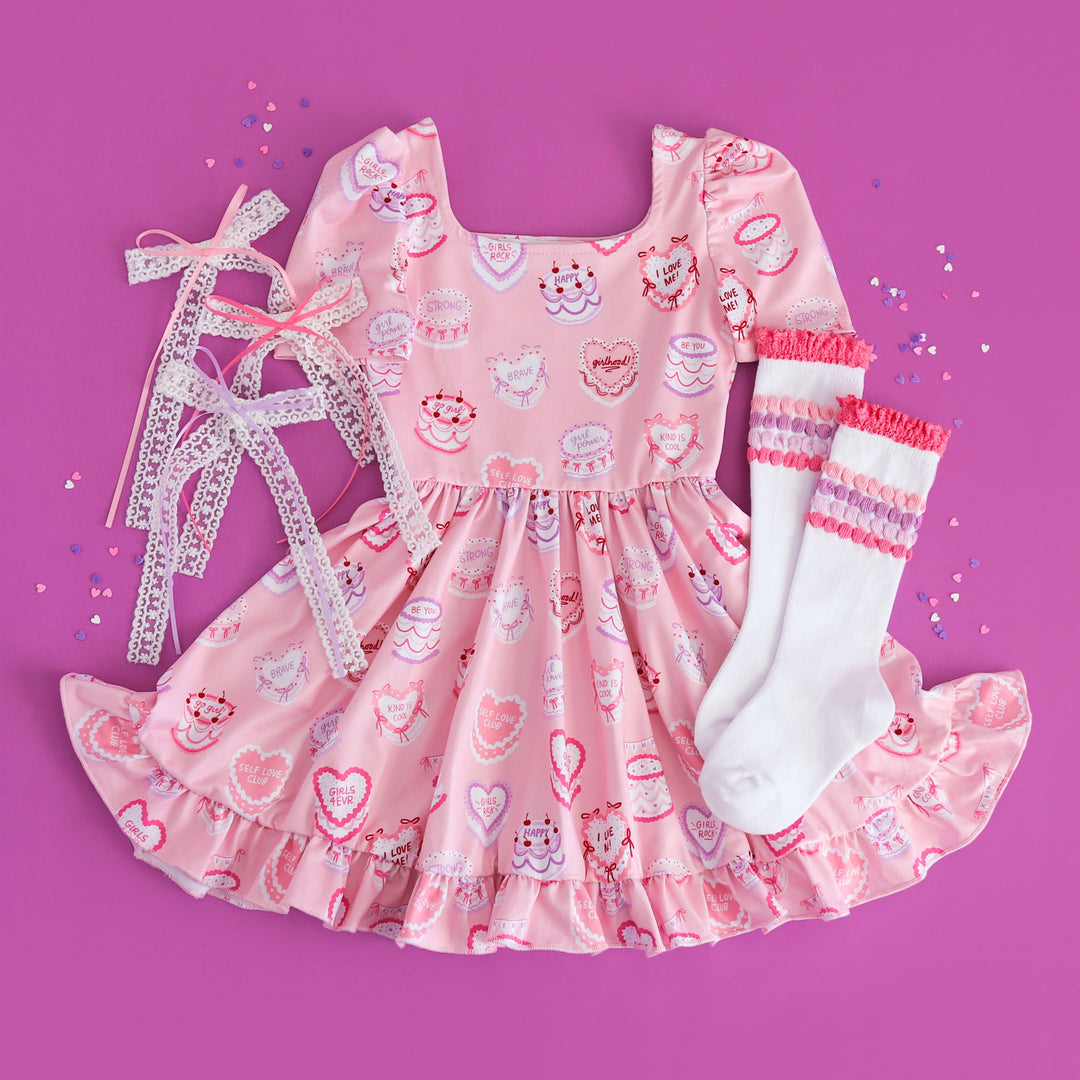 girls valentines flat lay styled with heart shaped cake featuring cake themed twirl dress, white knee high socks with pink and purple bubble accents and matching lace hair bows