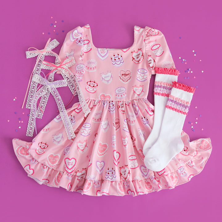 girls valentines flat lay styled with heart shaped cake featuring cake themed twirl dress, white knee high socks with pink and purple bubble accents and matching lace hair bows