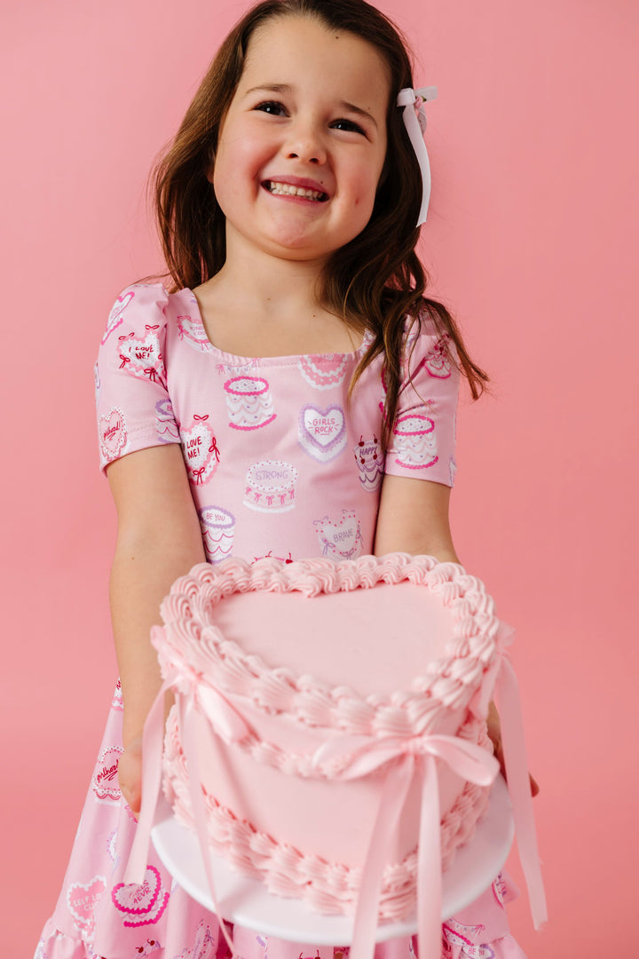 young girl holding heart shaped cake wearing pink cake print twirl dress decorated with words of affirmation for Valentine's day