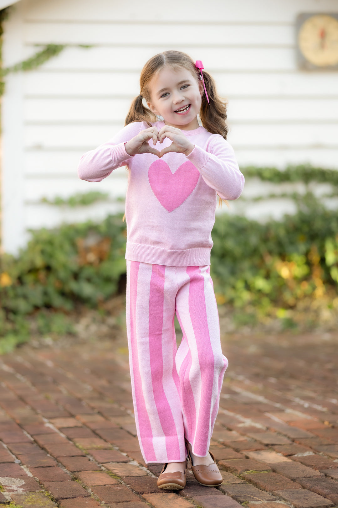 little girl with heart hands wearing two tone pink pullover sweater and striped two tone pink sweater pants for valentines day