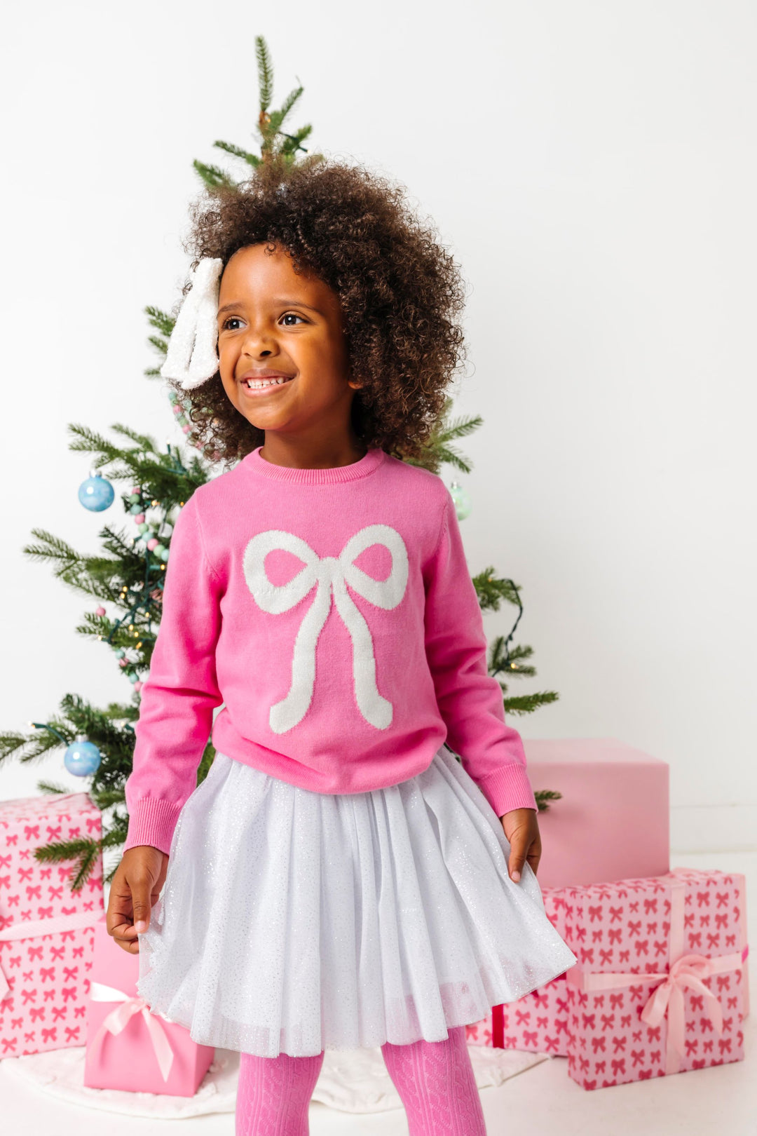 young girl in front of Christmas tree in pink cotton knit sweater with sparkly white bow design paired with white sparkly skirt and sequin hair bow