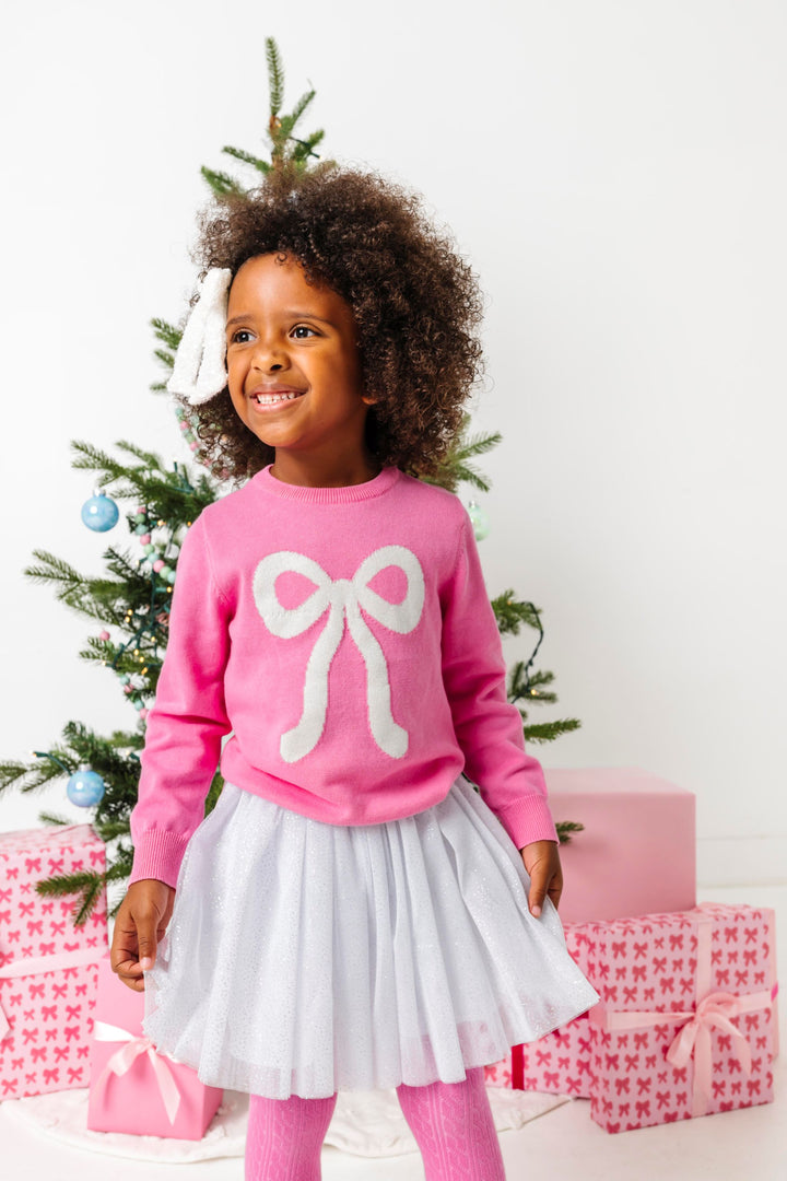 young girl in front of Christmas tree in pink cotton knit sweater with sparkly white bow design paired with white sparkly skirt and sequin hair bow
