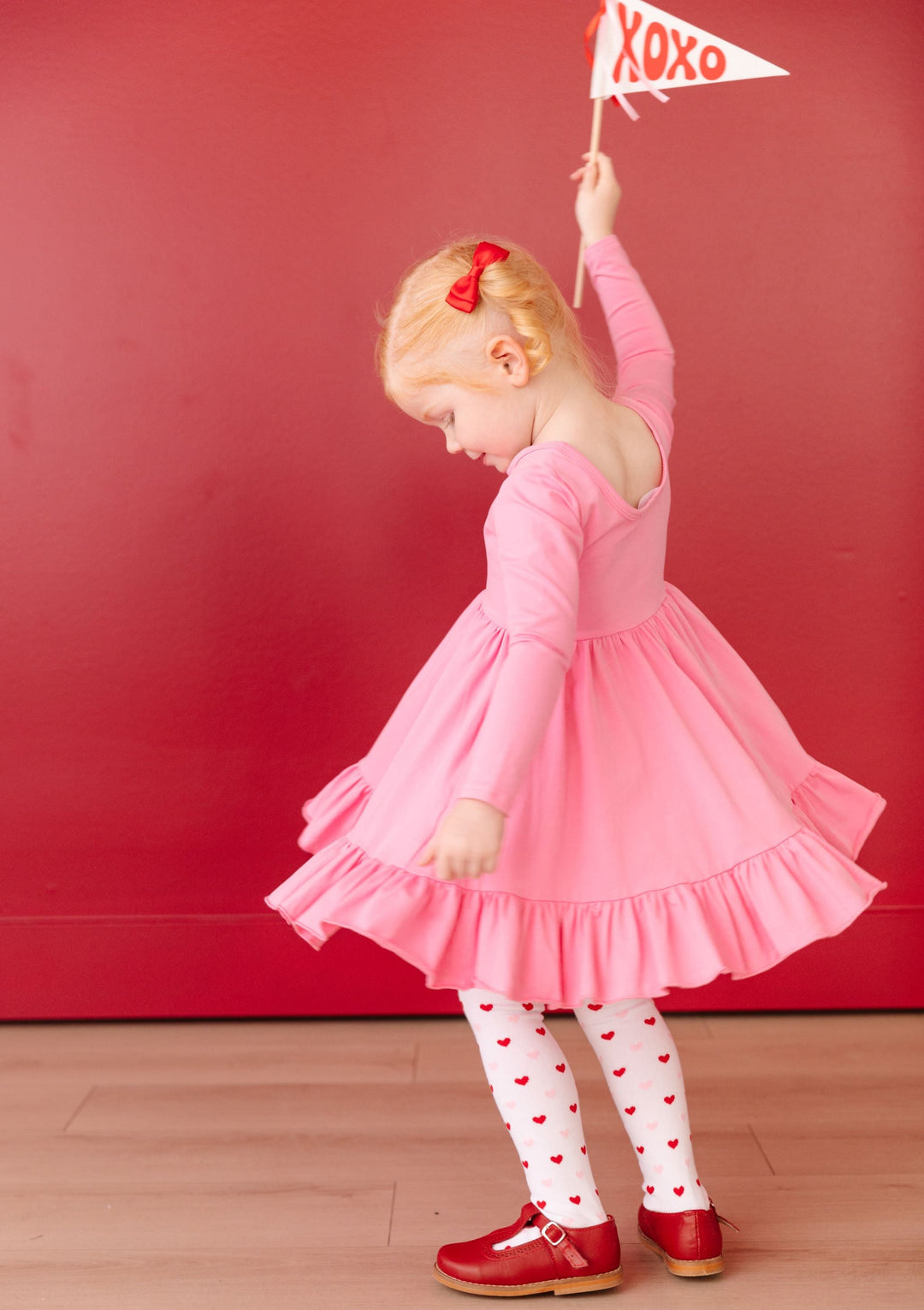 little girl twirling in long sleeve pink cotton dress with matching red and pink heart tights and red satin bows