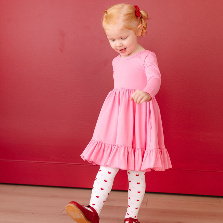 little girl in long sleeve pink cotton twirl dress with matching red and pink heart tights and red satin pigtail bows for Valentine's Day