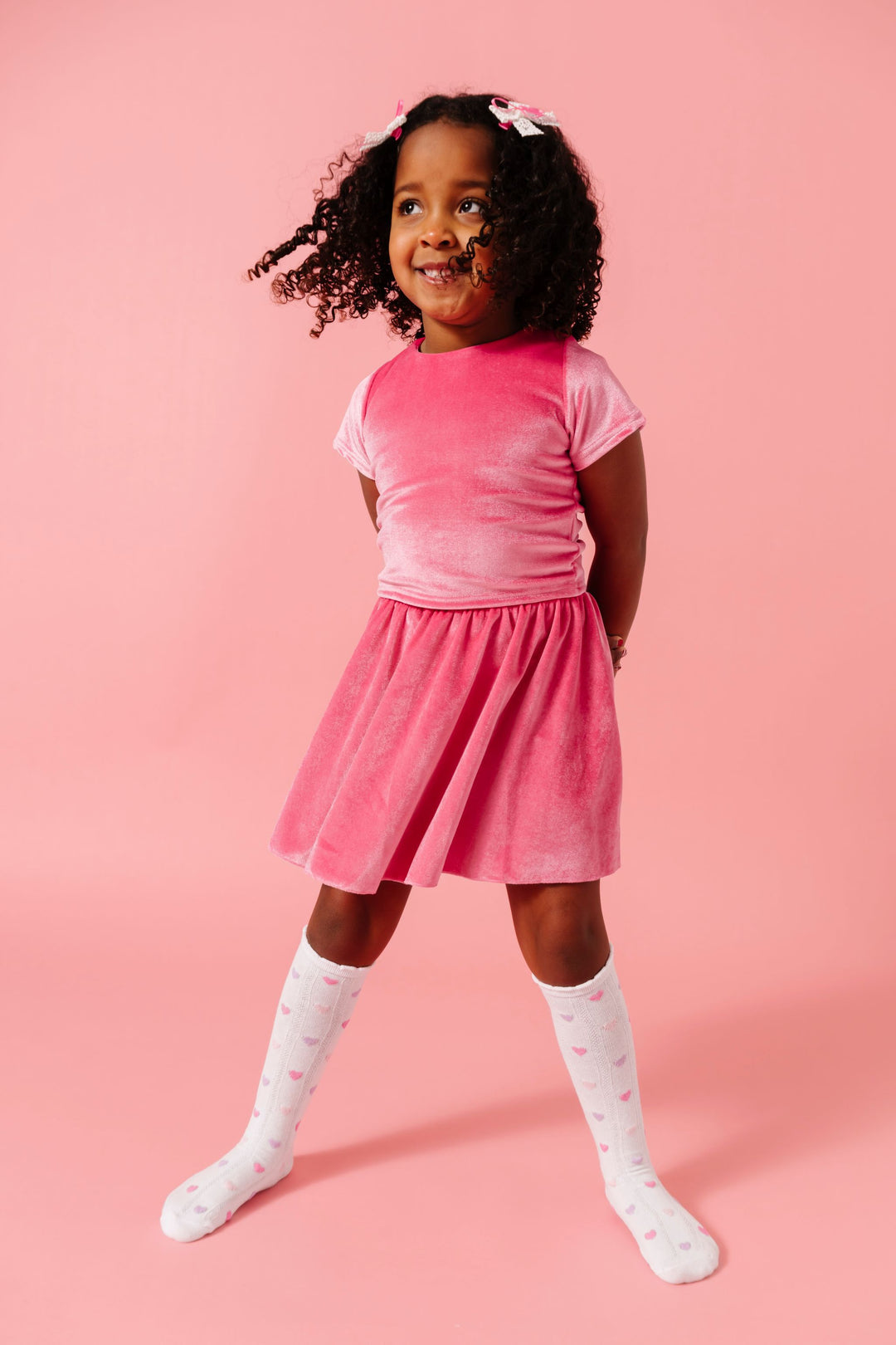 little girl in matching pink velvet fitted top and skort set with pastel heart knee high socks and matching white and pink lace ribbon pigtail bows