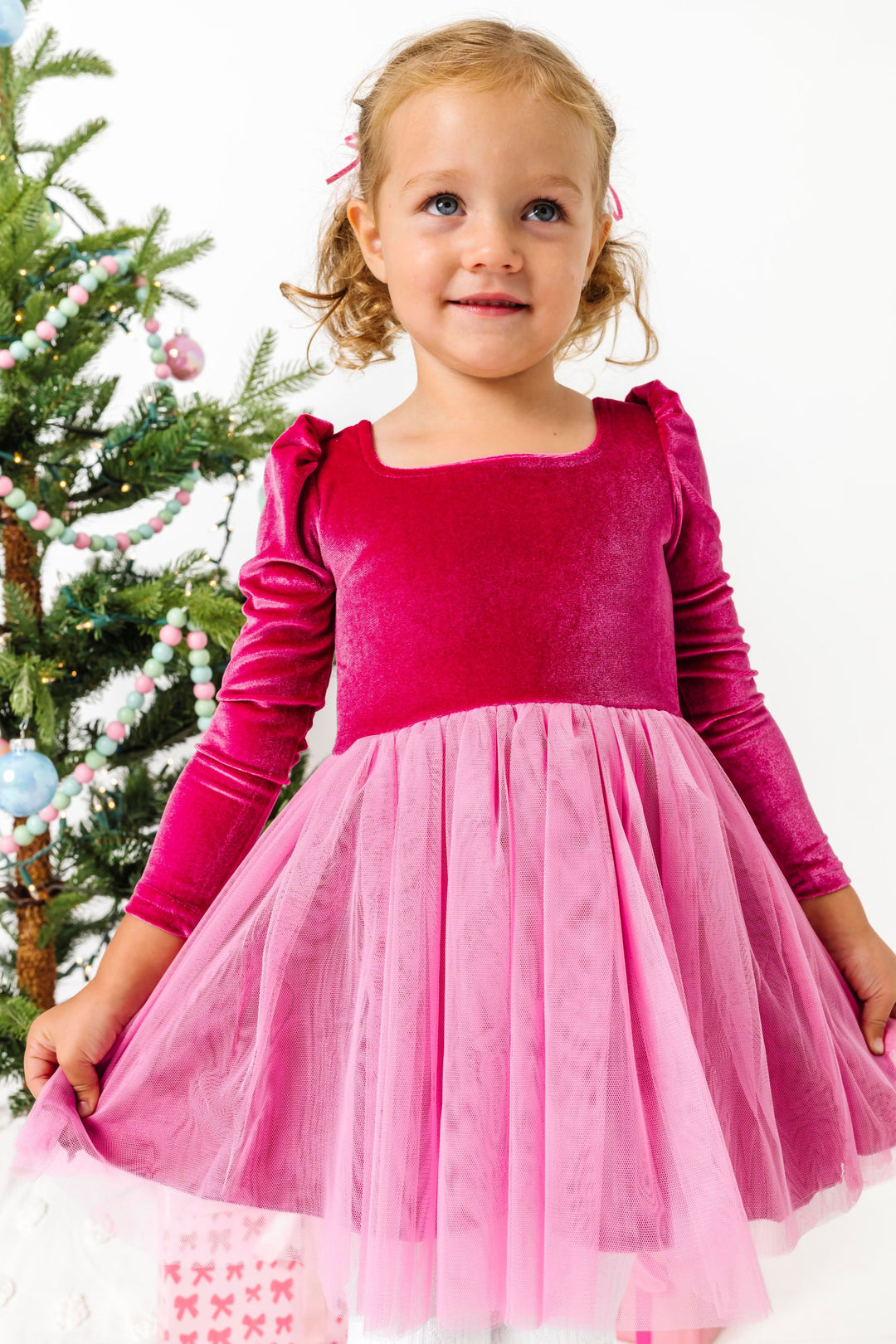 little girl by Christmas tree in bright pink velvet long sleeve dress with pink tulle skirt
