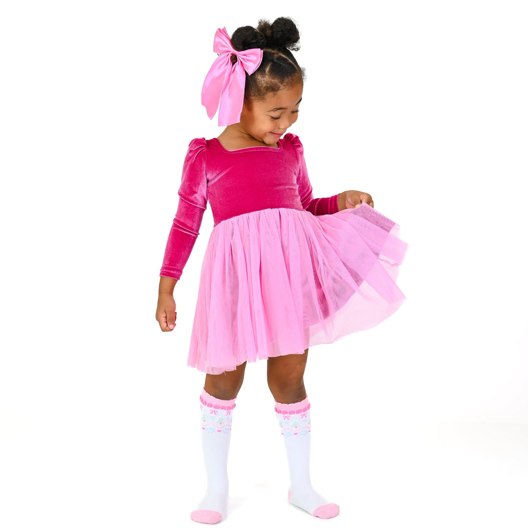 little girl in bright pink velvet long sleeve party dress with pink tulle knit paired with matching satin pink hair bow and white knee high socks with fair isle print