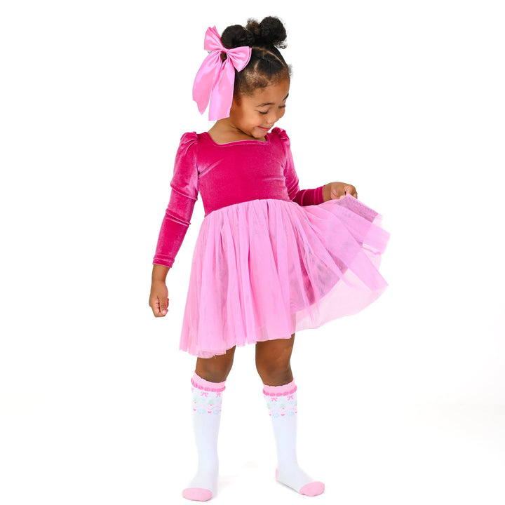 little girl in bright pink velvet long sleeve party dress with pink tulle knit paired with matching satin pink hair bow and white knee high socks with fair isle print
