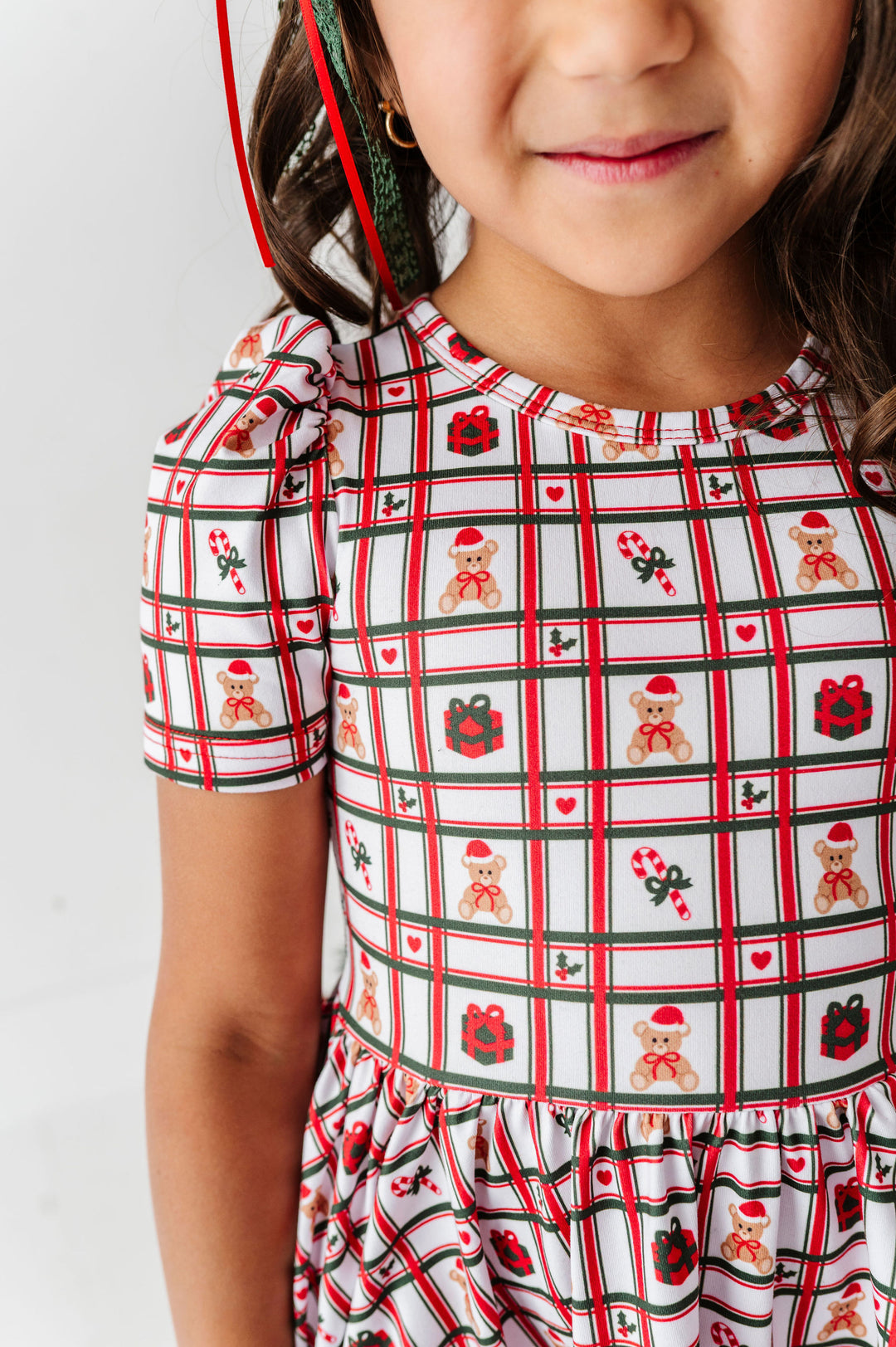 A close-up of a young girl wearing a short-sleeve dress with a festive holiday print. The dress features a white background with a red and green plaid grid pattern, decorated with teddy bears wearing Santa hats, red gifts, candy canes, and small red hearts.