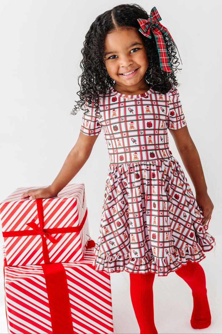 A young girl with curly black hair wearing a red, white, and black holiday plaid dress and red tights, standing next to a stack of wrapped Christmas presents. She is wearing a matching red and black plaid bow in her hair, set against a plain white studio background.