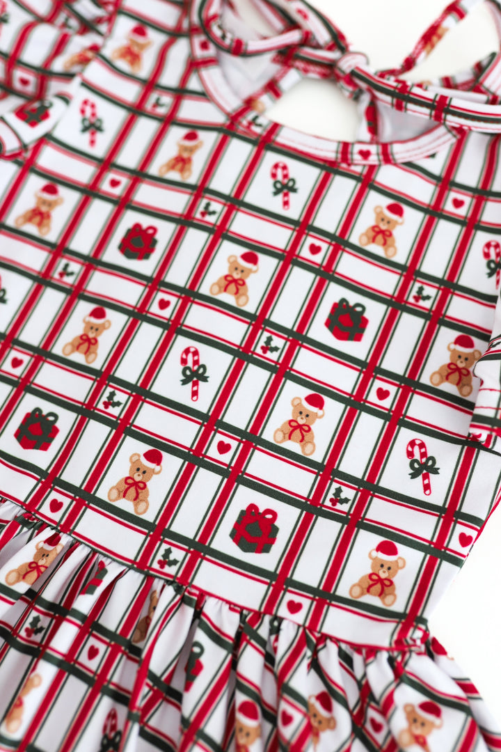 A close-up of a white holiday-themed plaid dress featuring teddy bears, candy canes, gifts, and red-and-black stripes, with a ruffled hem and tie-neck straps.