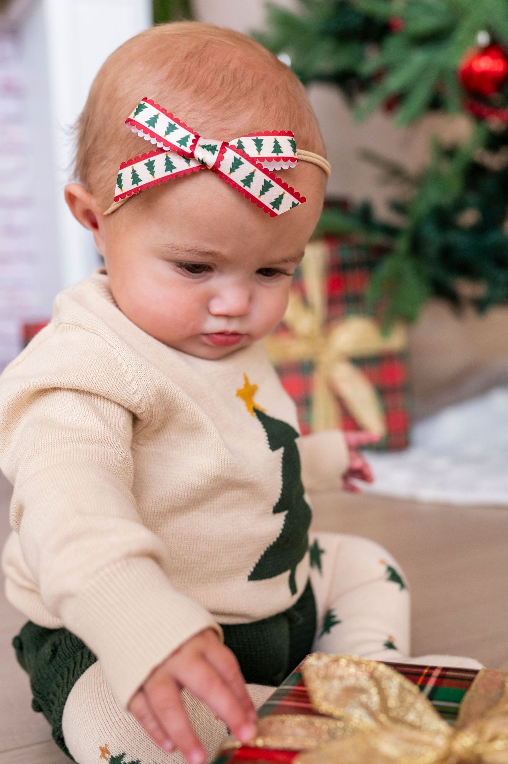 baby wearing xmas tree knit outfit with xmas tree ribbon hair bow touching wrapped xmas present