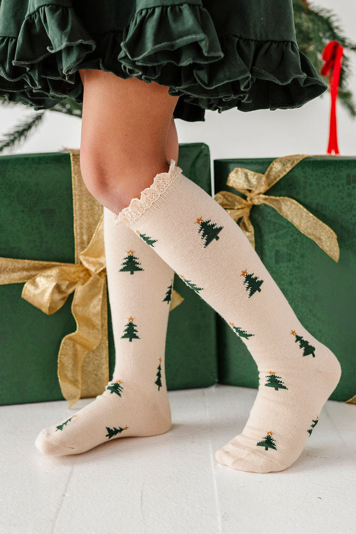 girl standing in front of christmas gifts wearing cream colored knee high socks with little green christmas trees on them and lace trim