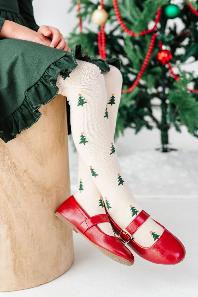 a little girl sitting in front of a christmas tree wearing cream colored knit tights with little star topped christmas trees on them