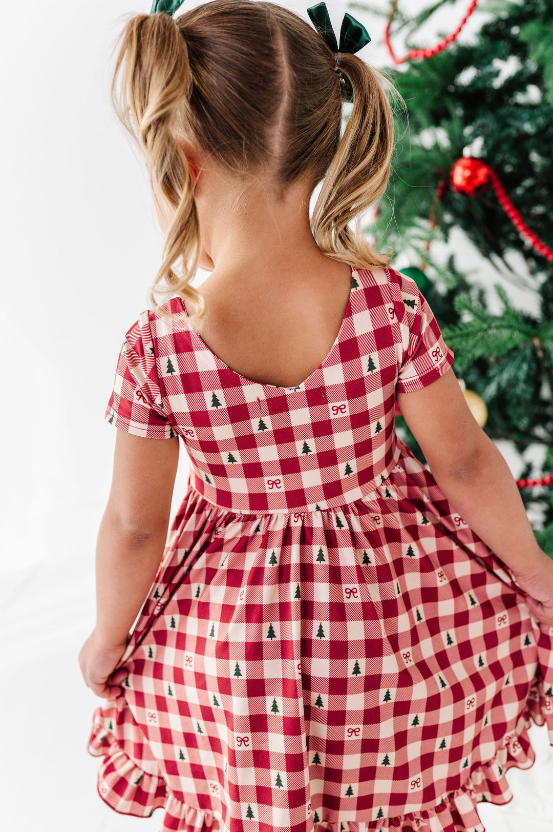 A young girl viewed from behind wearing a red and white gingham dress with a small Christmas tree print, standing next to a decorated Christmas tree.