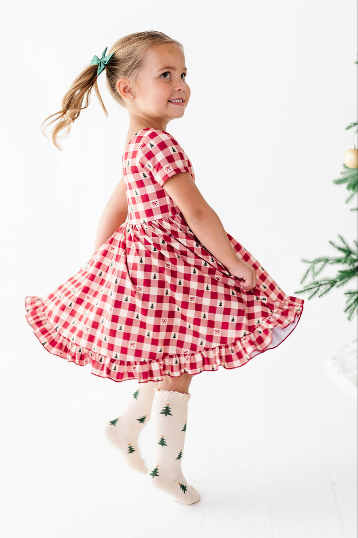 A young girl with blonde hair in a ponytail wearing a red and white gingham dress and Christmas tree print socks, captured mid-spin in a bright white studio setting.