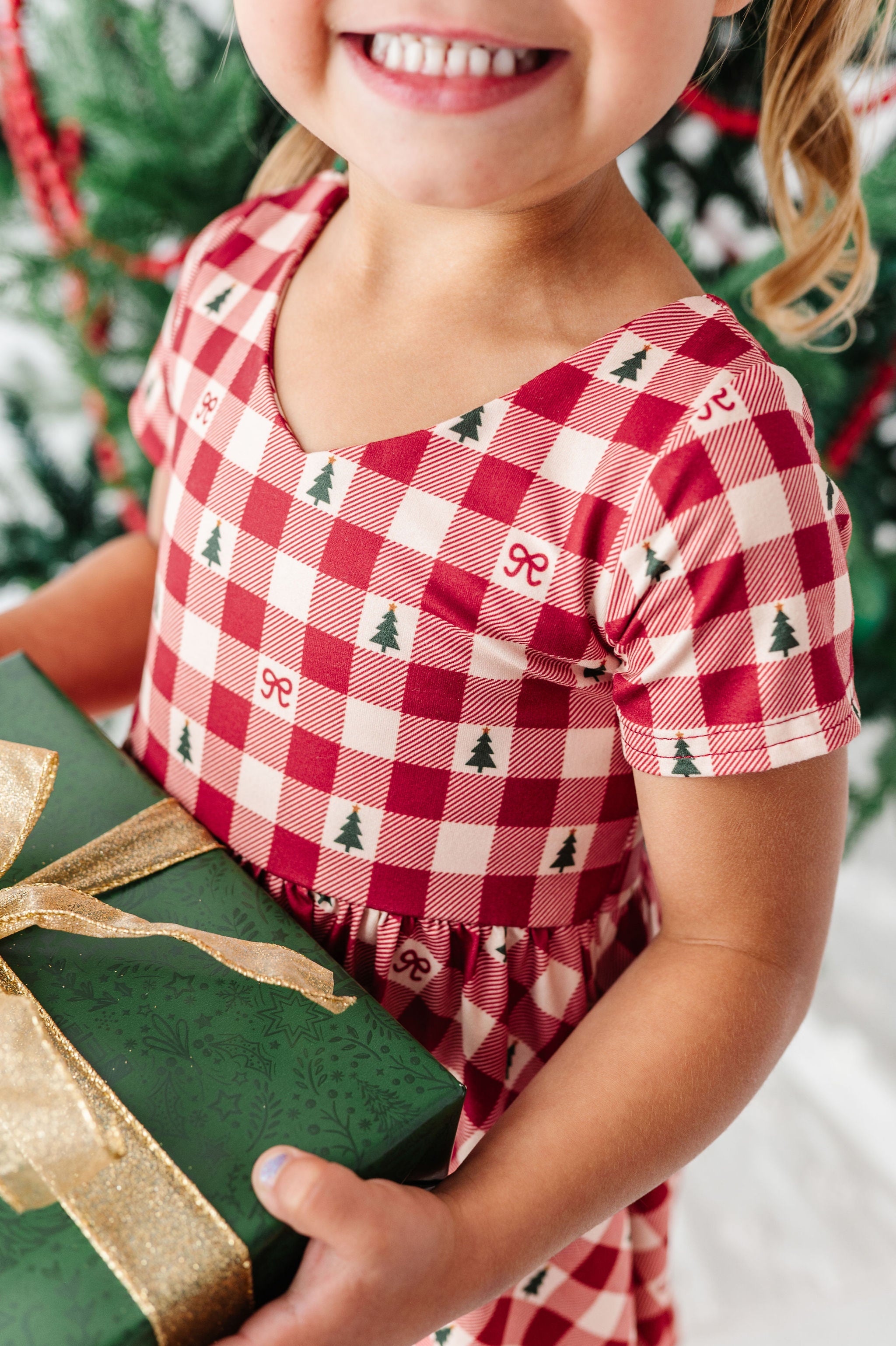 A close-up of a smiling child wearing a red and white gingham dress with small green Christmas tree and bow motifs, holding a dark green gift box with a gold ribbon in front of a blurred Christmas tree background.