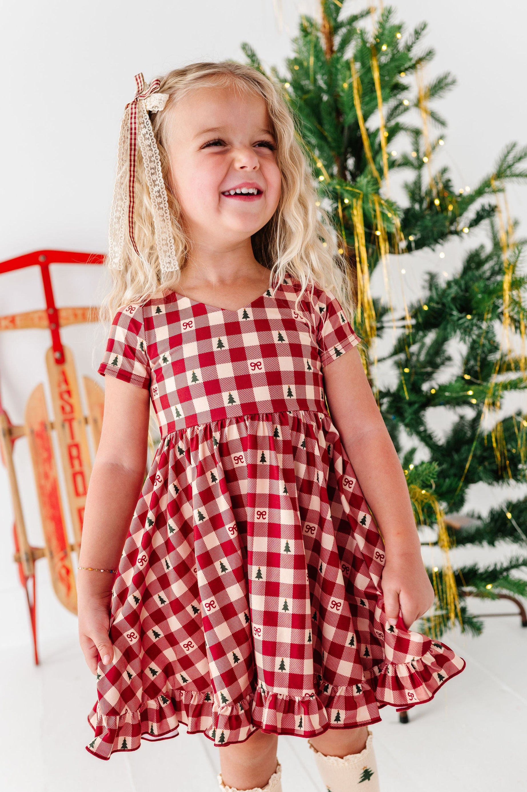 A young girl with blonde hair wearing a red and white gingham dress with a Christmas tree pattern, standing in front of a decorated Christmas tree and a red sled.