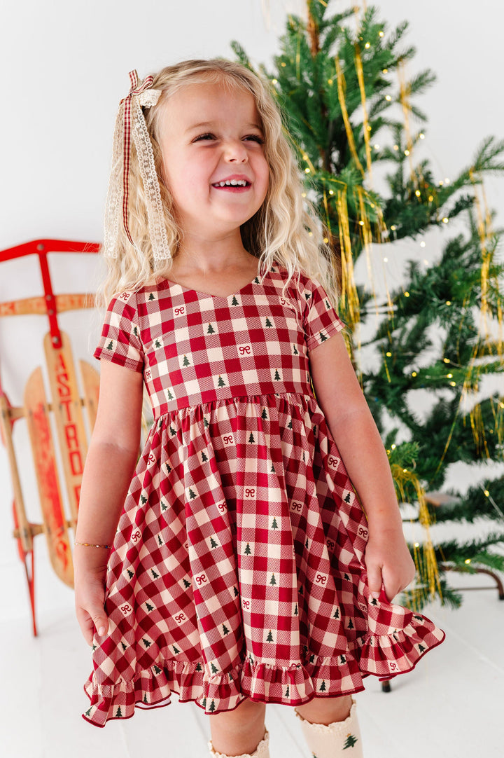 A young girl with blonde hair wearing a red and white gingham dress with a Christmas tree pattern, standing in front of a decorated Christmas tree and a red sled.