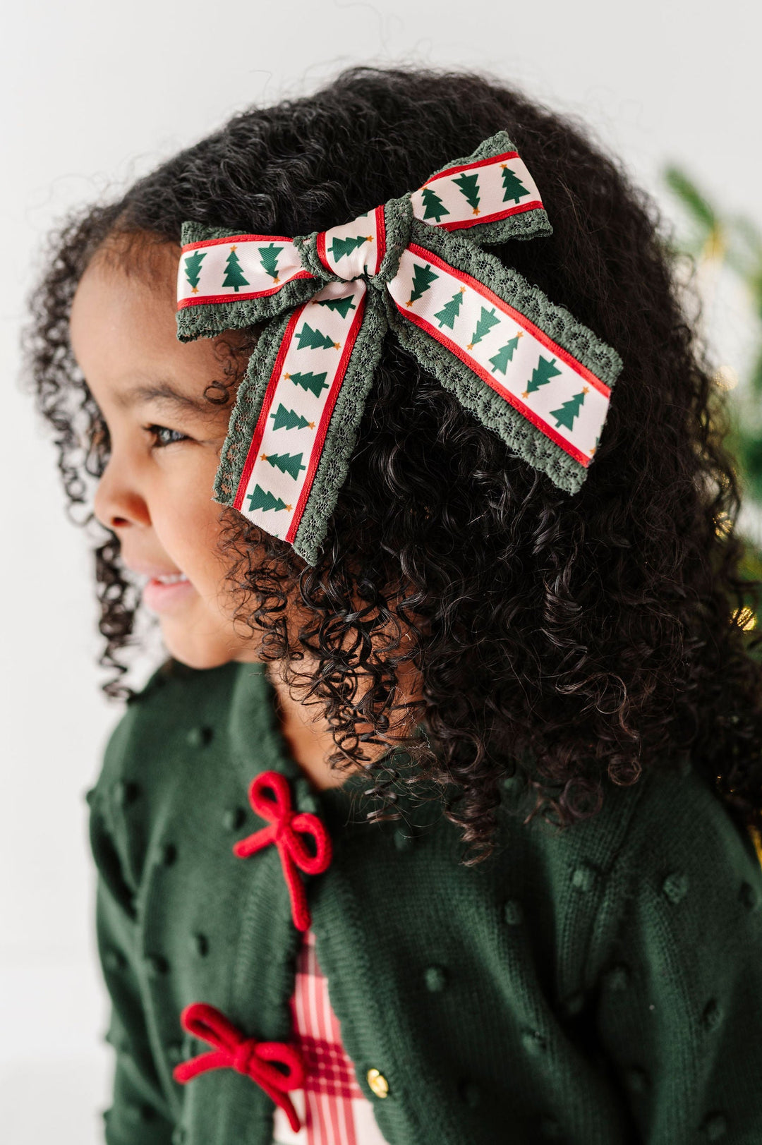 A young child with curly dark hair wearing a festive green cardigan with red bow details and a plaid top, wearing a large patterned hair bow. The background is a soft white with a blurred Christmas tree on the right.