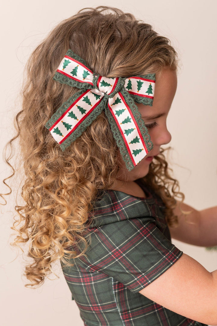 A young girl with curly blonde hair wearing a green plaid dress and a matching green, cream, and red xmas tree ribbon hair bow, decorating a Christmas tree.