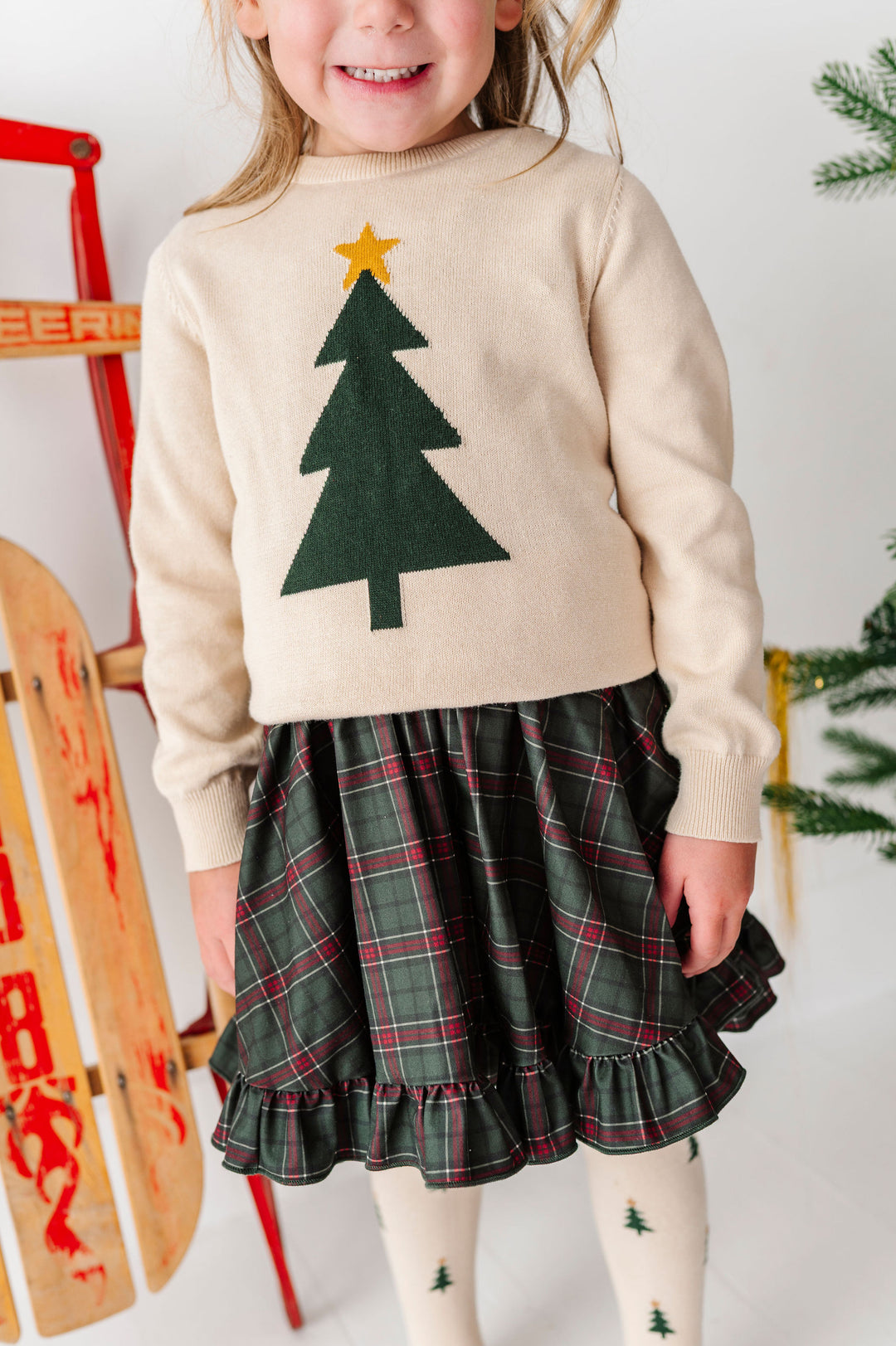 A young girl wearing a cream knit sweater with a green felt Christmas tree and gold star, paired with a green plaid ruffled skirt and white tights with small tree prints.
