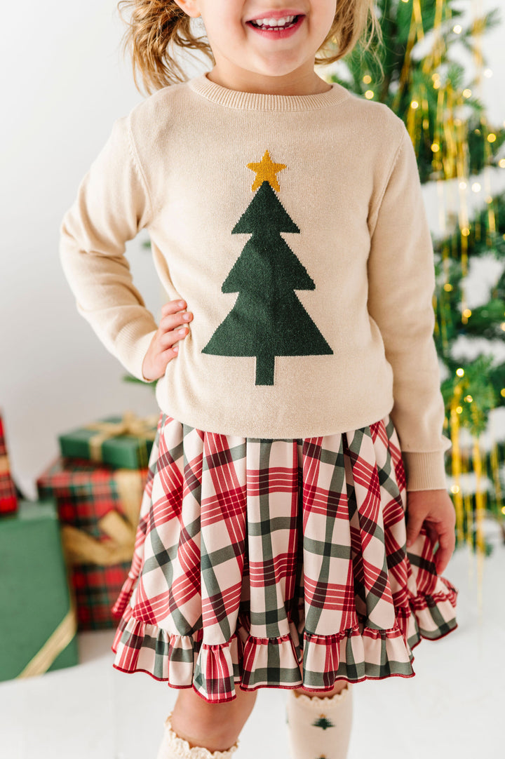 A young child wearing a cream knit sweater with a green felt Christmas tree and gold star, paired with a red, green, and white plaid ruffled skirt, standing in front of a decorated Christmas tree and wrapped gifts.