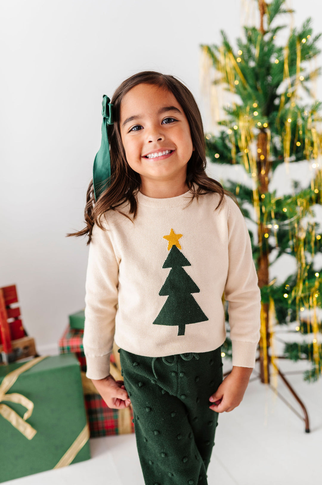 A young girl smiling in a cream long-sleeve sweater with a green felt Christmas tree applique and a gold star, paired with dark green pants, standing in front of a decorated Christmas tree.