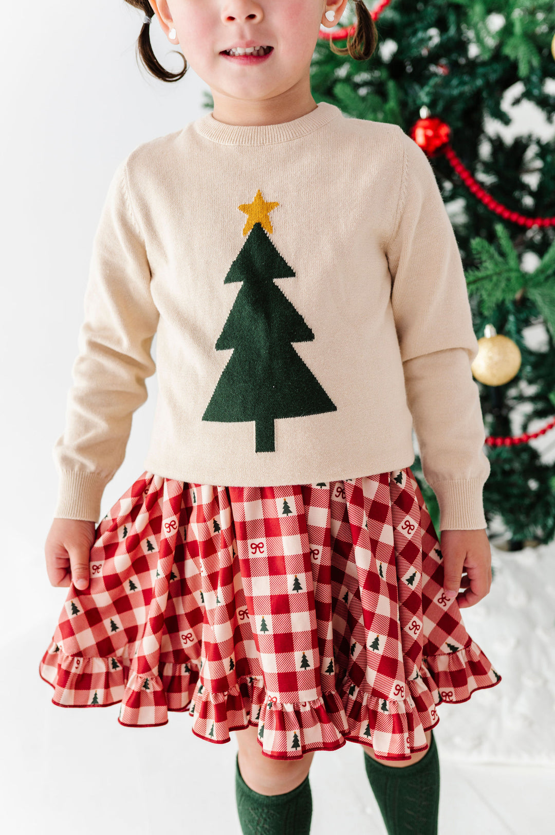 A young child wearing a cream knit sweater with a green felt Christmas tree and gold star, paired with a red and white gingham ruffled skirt with small tree prints, standing in front of a decorated Christmas tree.