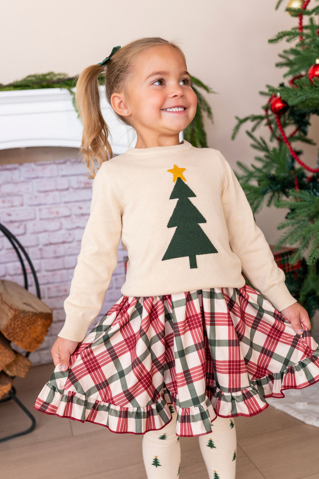 A young girl with blonde pigtails stands indoors wearing a cream knit sweater with a green Christmas tree and yellow star, paired with a red, green, and cream plaid ruffled skirt and matching cream tights. A decorated Christmas tree and fireplace are visible in the background.