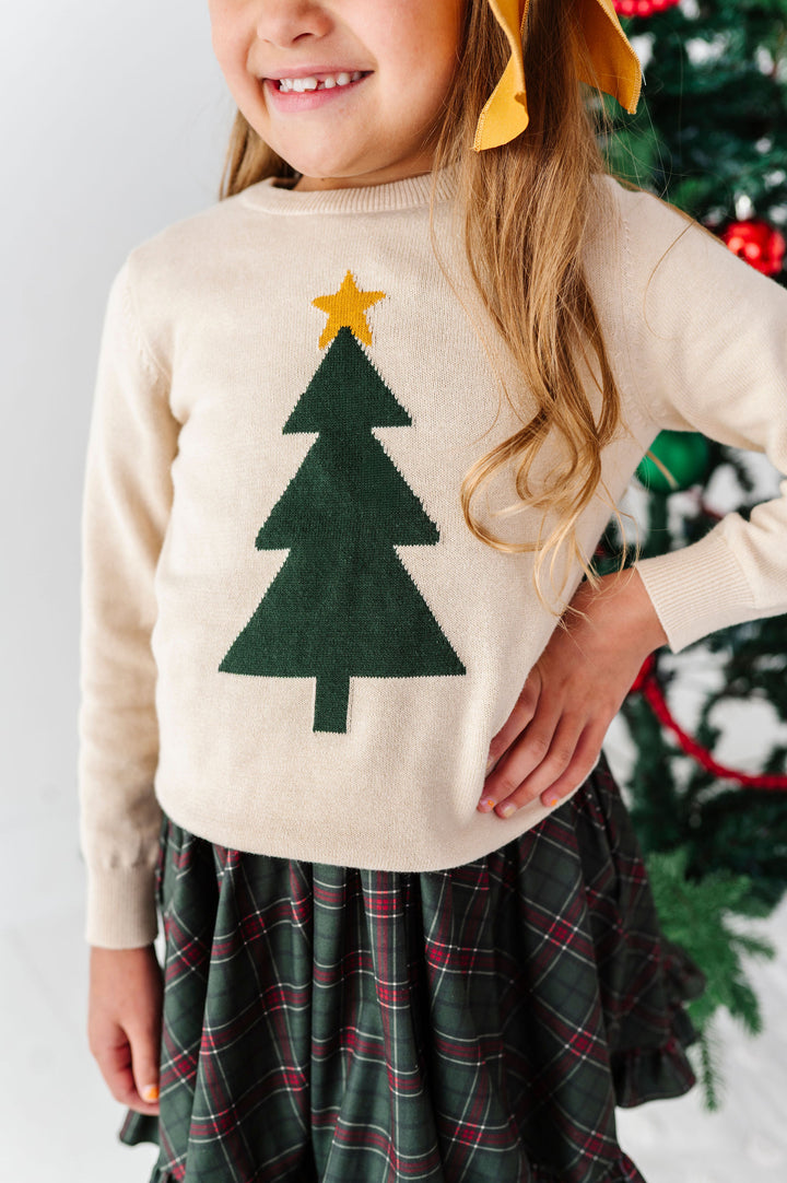 A child wearing a cream knit sweater with a green Christmas tree and yellow star motif, paired with a green plaid skirt, standing in front of a decorated Christmas tree.