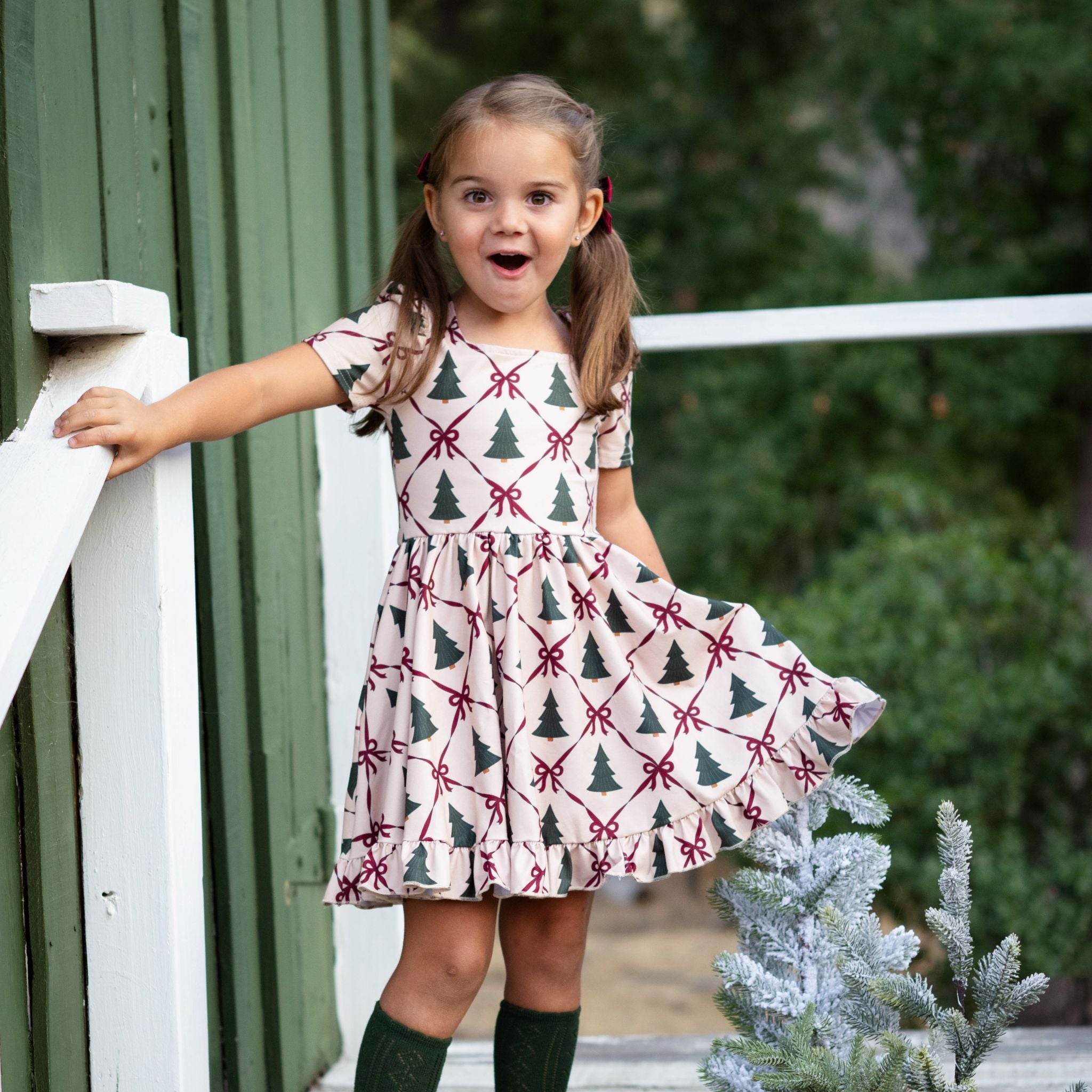 little girl at tree farm in cream twirl dress with tree and crimson ribbon print paired with forest green open knit knee high socks