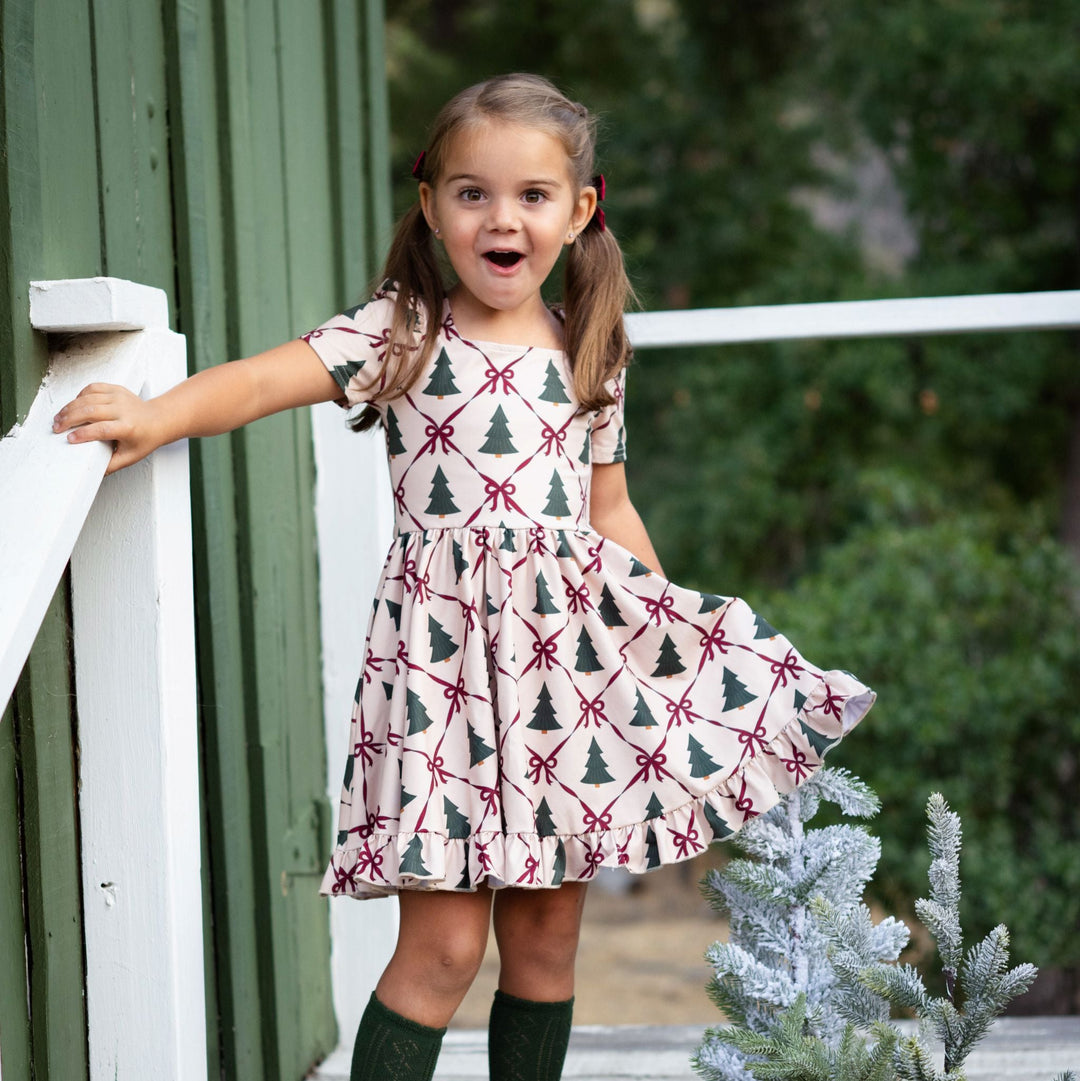 little girl at tree farm in cream twirl dress with tree and crimson ribbon print paired with forest green open knit knee high socks
