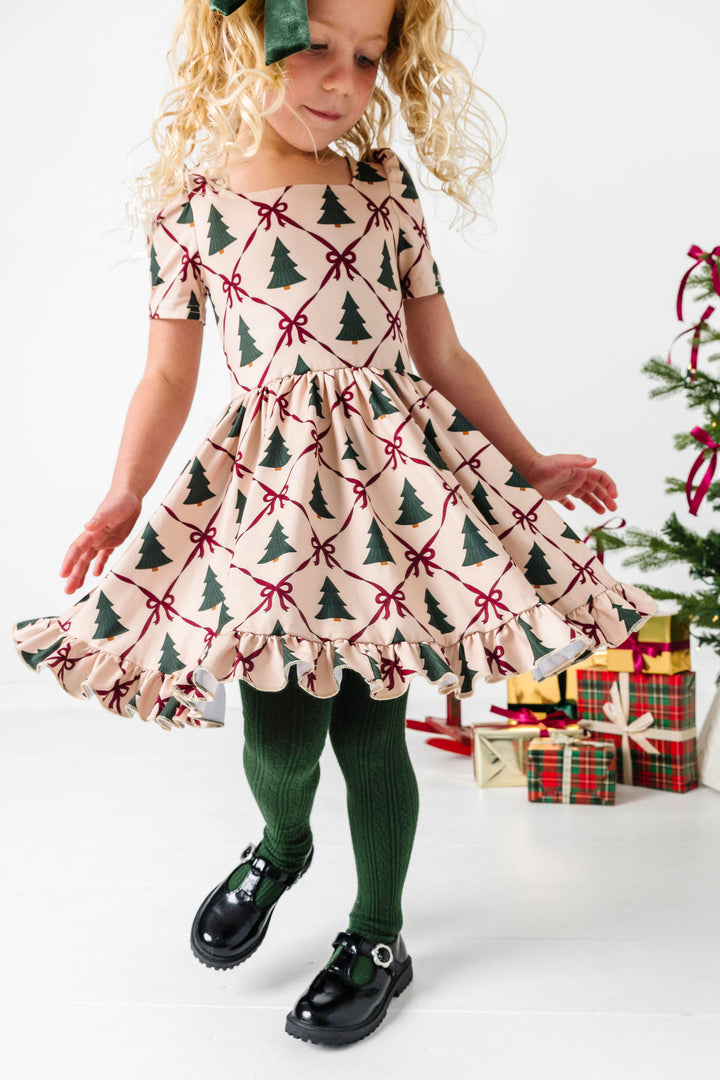 girl twirling in front of Christmas presents in cream short sleeve twirl dress with crimson bow and forest tree print paired with matching green cable knit tights