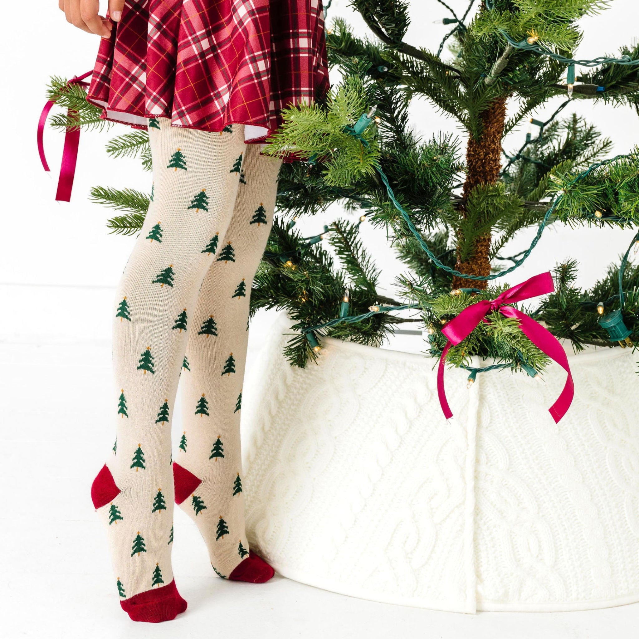 little girl on tip toes decorating tree in cream tights with Christmas tree pattern and crimson plaid outfit