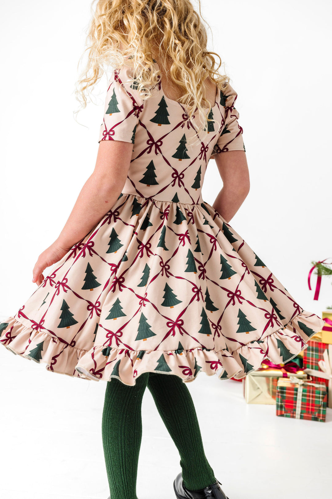 girl twirling in front of Christmas presents in cream short sleeve twirl dress with crimson bow and forest tree print paired with matching green cable knit tights