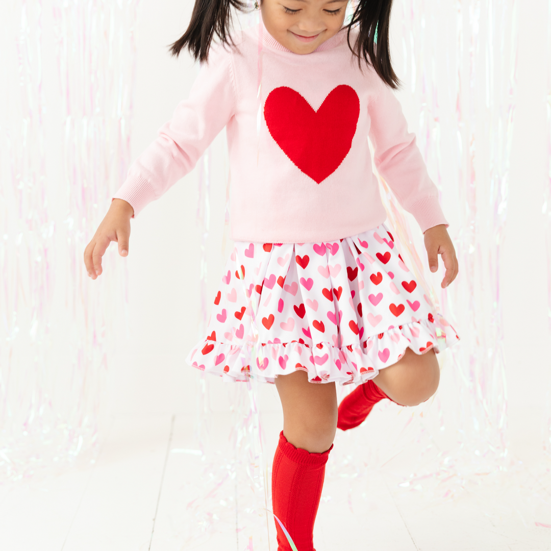 girl skipping in white dress with red and pink heart print and pink sweater with red heart and bright red knee highs