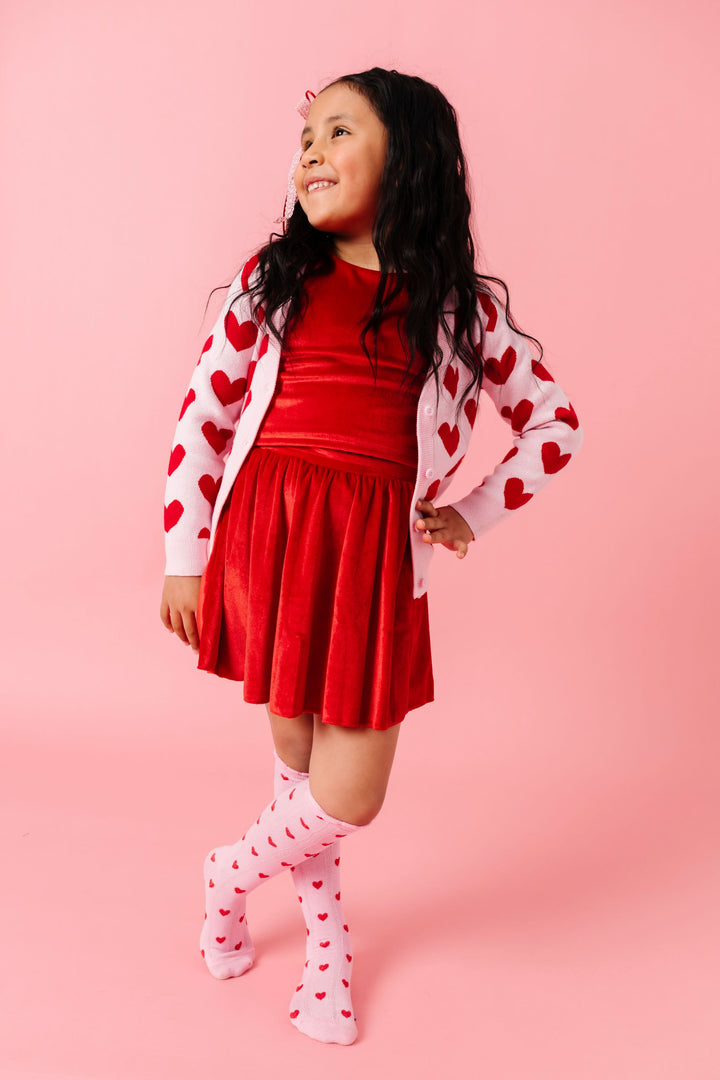 young girl wearing matching bright red velvet top and skort set paired with pink knit cardigan with red heart design and made to match heart knee socks