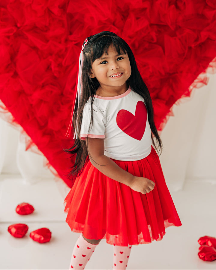 little girl in front of red heart backdrop wearing top with red heart design, bright red tulle skirt and light pink socks with red hearts