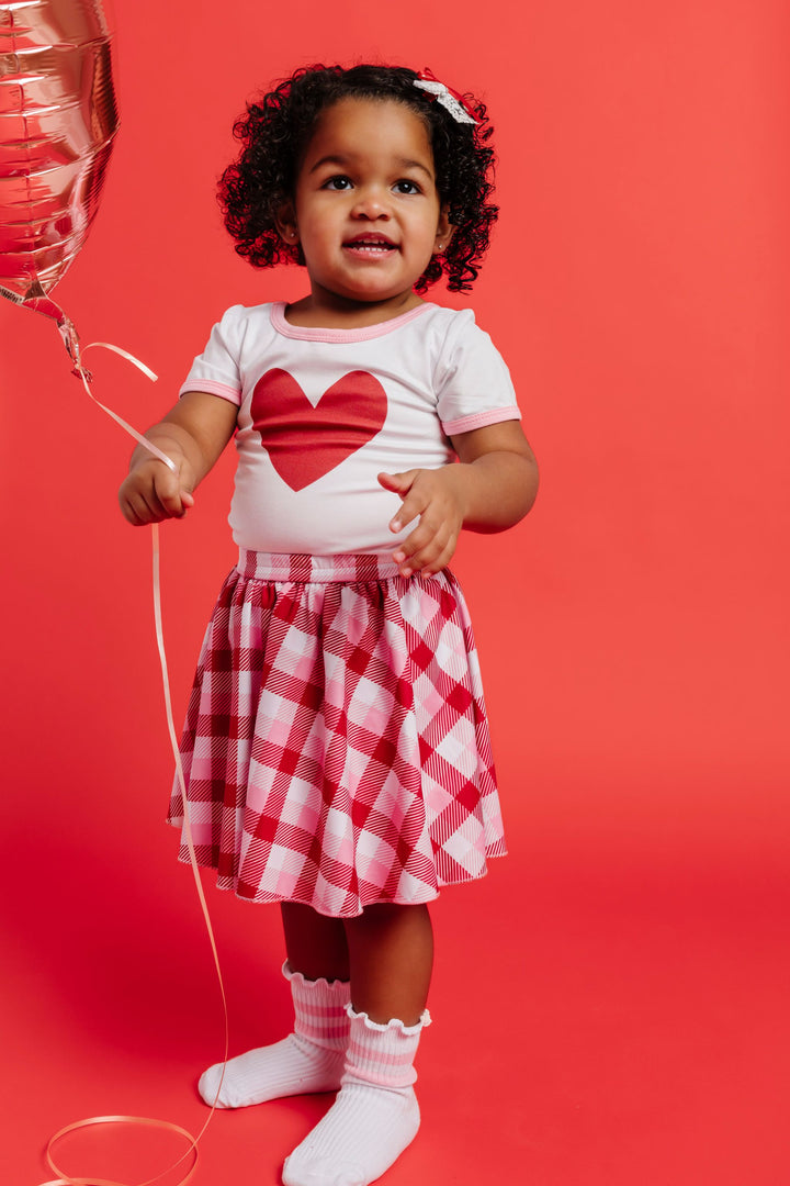 little girl holding heart balloon in white bamboo top with red heart and plaid valentines skort with ribbed white midi socks