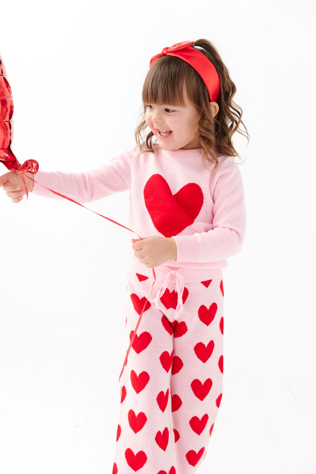 little girl holding balloon string in pink pullover sweater with red heart design and matching pink knit sweater pants with red heart pattern