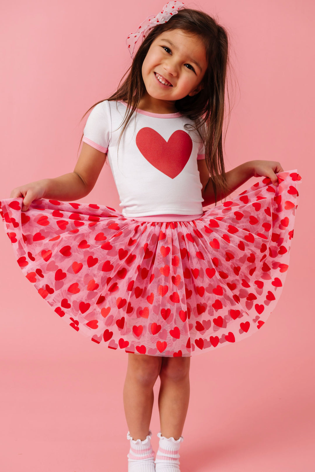 young girl wearing pink tutu skirt with red heart design paired with white fitted top with single red heart and pink trim
