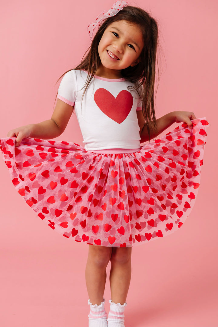 young girl wearing pink tutu skirt with red heart design paired with white fitted top with single red heart and pink trim