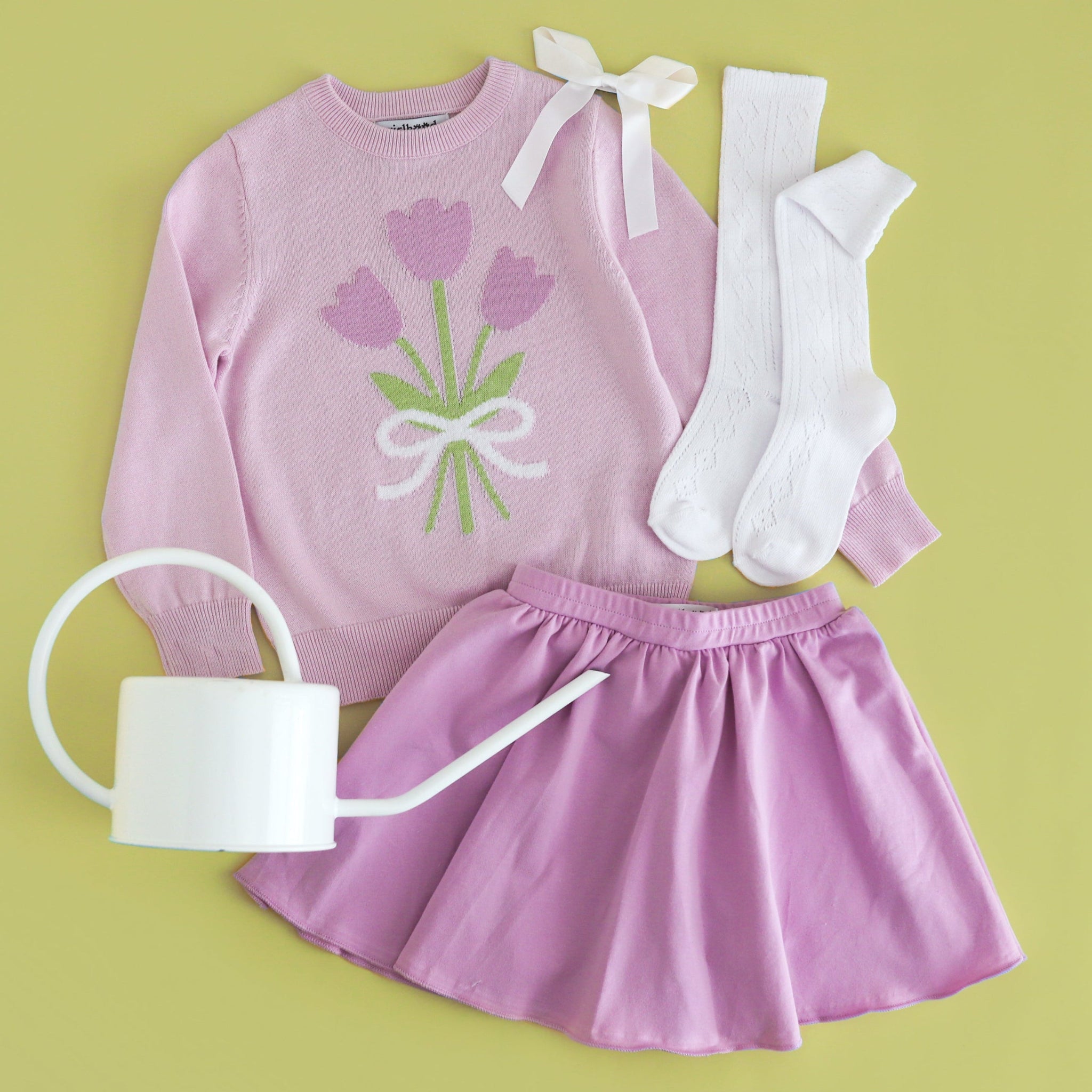 Children's outfit with pink top and skirt, white socks, and white watering can on a green background