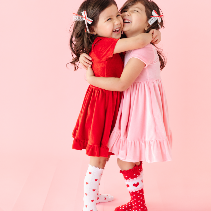 twin girls hugging wearing pink and red velvet valentine's day dresses and heart knee highs