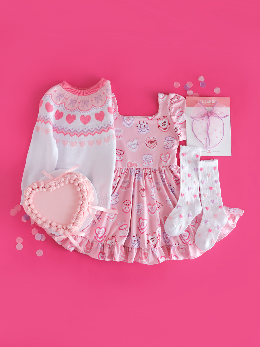 girls valentines flat lay styled with heart shaped cake featuring cake themed twirl dress, pastel Valentine's fair isle sweater, pastel heart socks and matching sheer bow