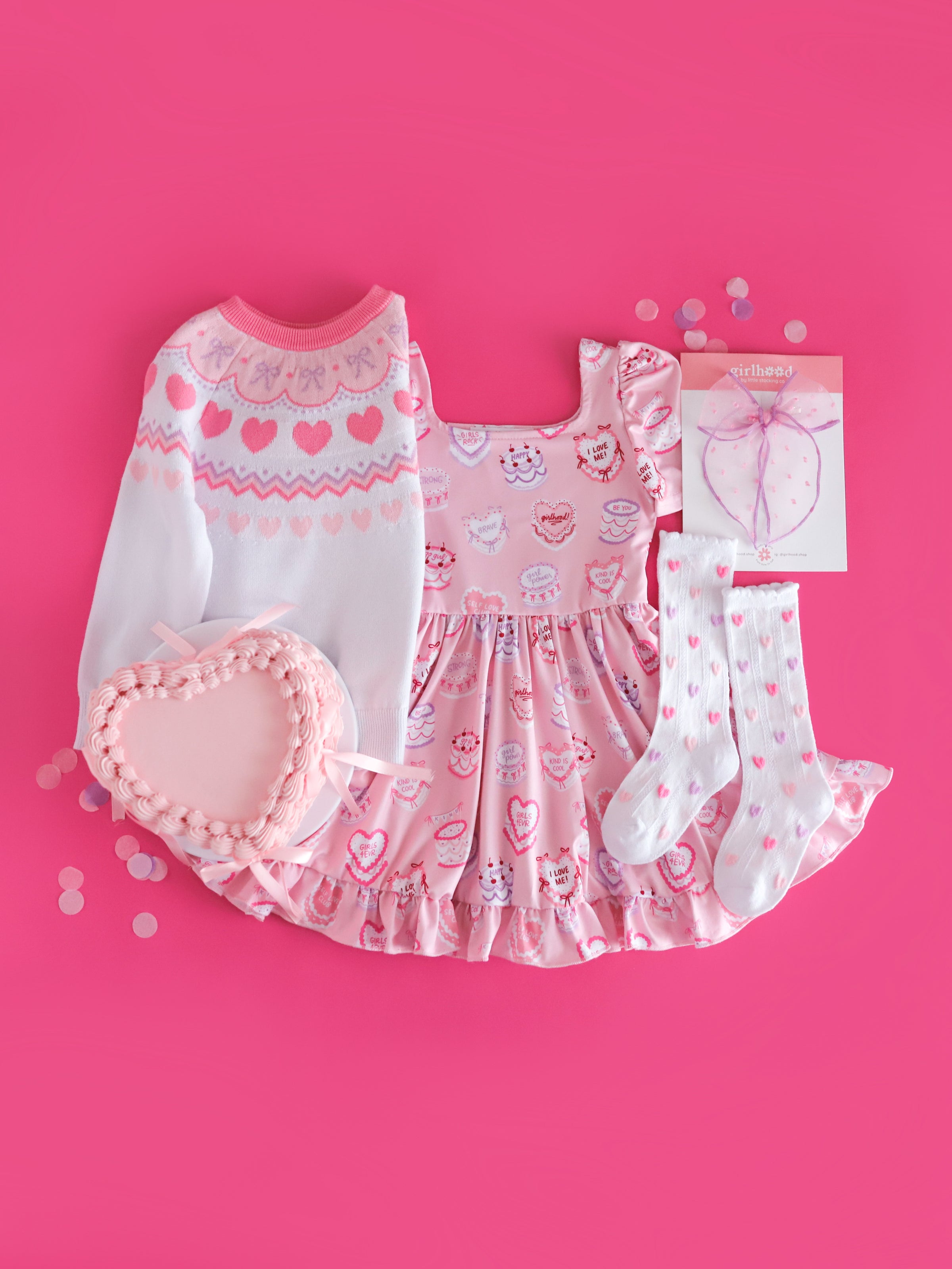 girls valentines flat lay styled with heart shaped cake featuring cake themed twirl dress, pastel Valentine's fair isle sweater, pastel heart socks and matching sheer bow