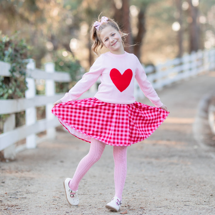 girl posing in valentines outfit of red and pink gingham dress and heart sweater with fancy knit pink tights